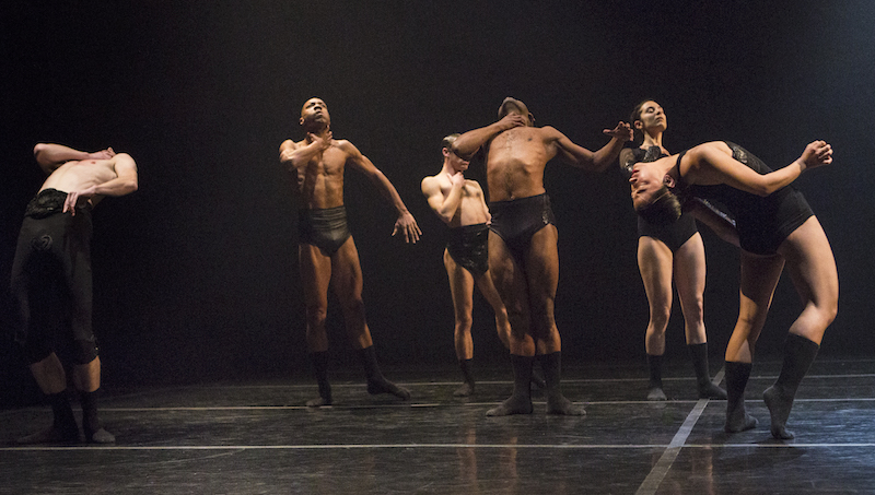 Dancers wearing black briefs stumble behind themselves with their hands around their necks. Two dancers perform deep back bends.